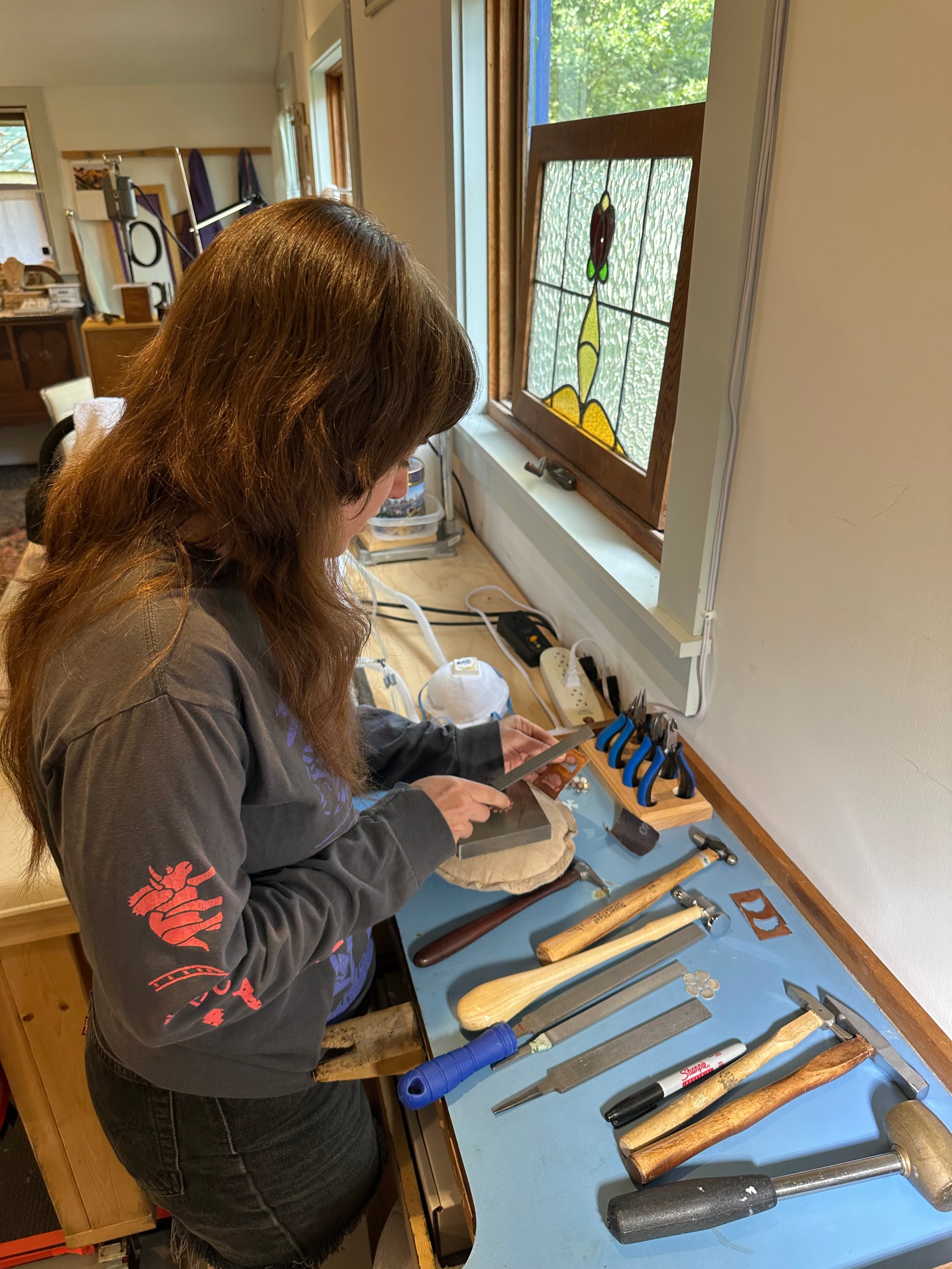 Person working on a project at a workbench with tools and a stained glass window in the background.